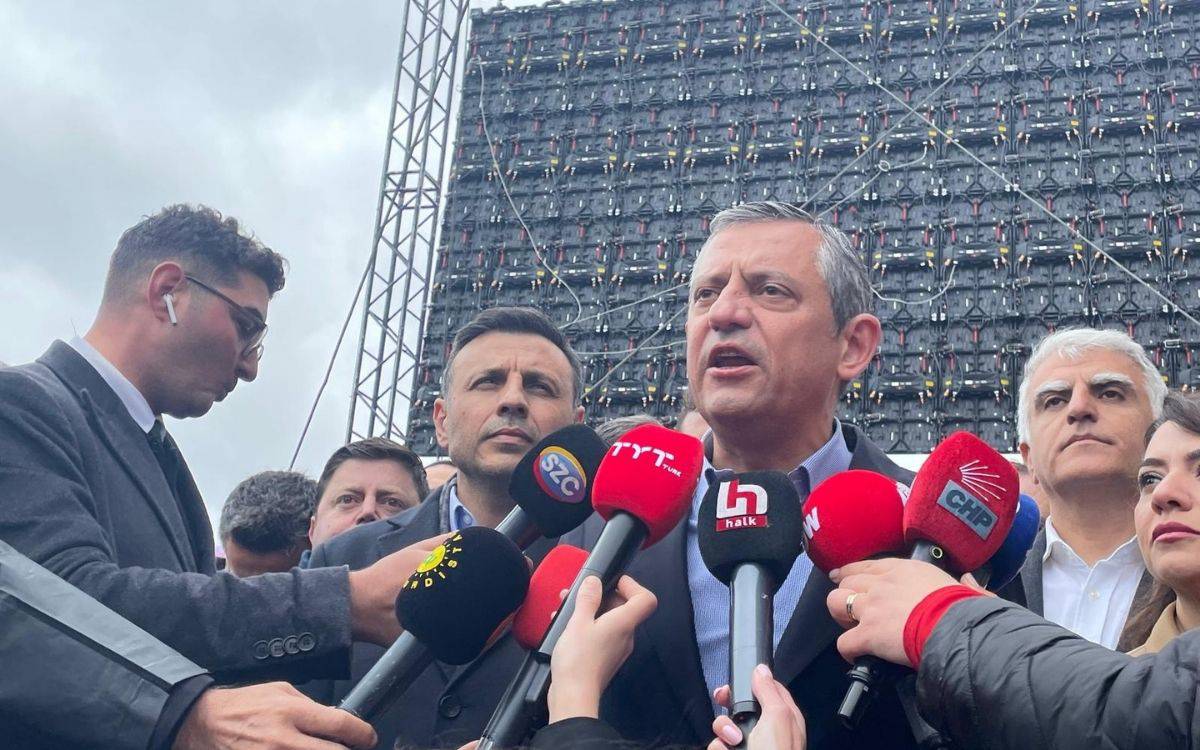 Fotoğraf, kalabalık bir basın açıklaması anını yansıtıyor. Görselin merkezinde CHP Genel Başkanı Özgür Özel, çok sayıda mikrofonun önünde konuşma yaparken görülüyor. Yüz ifadesi ciddi ve kararlı; dudakları açık, konuşma anında. E