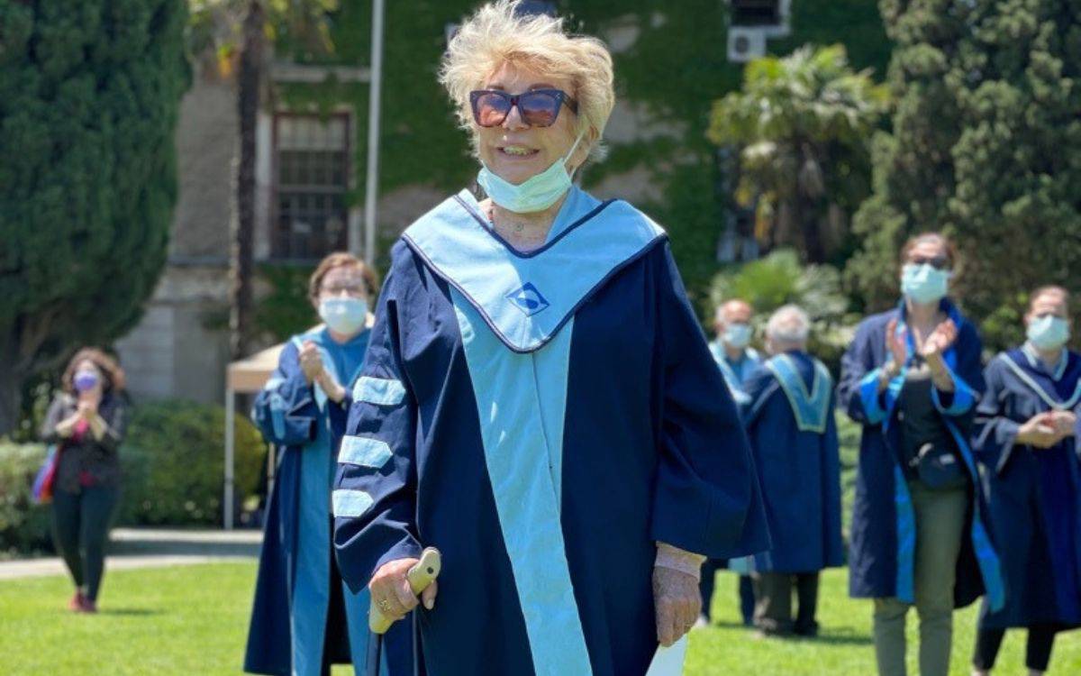 Fotoğrafta, Prof. Dr. Oya Başak, Boğaziçi Üniversitesi kampüsünde, tören kıyafetiyle görülüyor. Üzerinde mavi tonlarında cübbe bulunan Başak, elinde bir belge veya telefon tutuyor. Gözlük takıyor ve yüzünde zarif bir tebessüm var. Boynundaki tıbbi maske çenesinin altına indirilmiş durumda.  Arka planda ise benzer cübbe giymiş, maske takan akademisyenler alkışlarla onu onurlandırıyor. Oya Başak çimlik bir alanda, muhtemelen açık hava bir tören sırasında yürüyor. Fotoğraf, hem akademik saygıyı hem de ona duyulan sevgiyi yansıtan, onurlu bir veda veya kutlama anını ölümsüzleştirmiş.